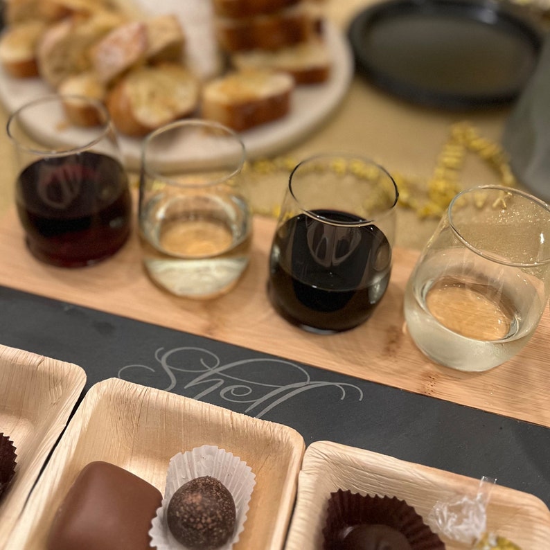 May include: A wooden serving board with four stemless wine glasses filled with red and white wine. Small square wooden plates hold chocolates. Sliced bread is in the background. The board has the word "Shell" etched on it.