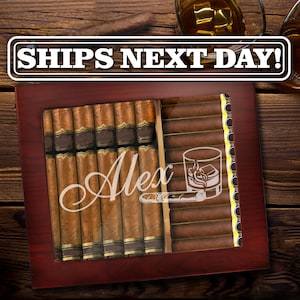 May include: A dark wood cigar humidor with a glass top featuring the name Alex in white script, a graphic of a glass of whiskey with ice, and a cigar. The humidor is filled with rows of brown cigars with gold bands, and the text SHIPS NEXT DAY!