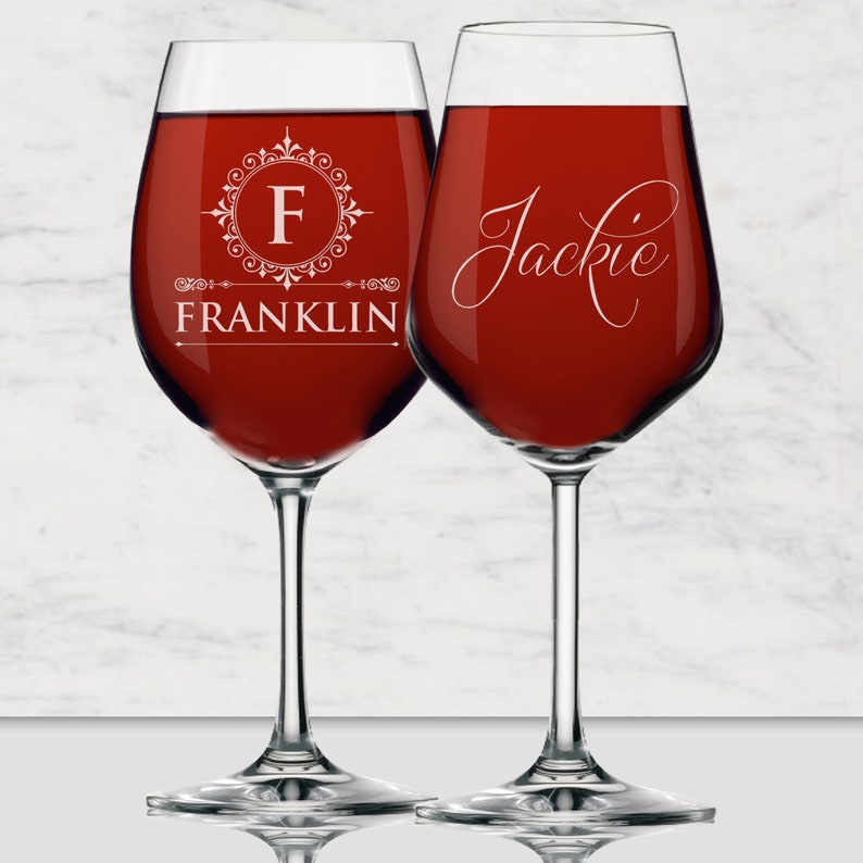 May include: Two clear glass wine glasses filled with deep red liquid. The left glass features a white circular filigree emblem with the letter F and the name FRANKLIN below. The right glass displays the name Jackie in a white cursive script font.