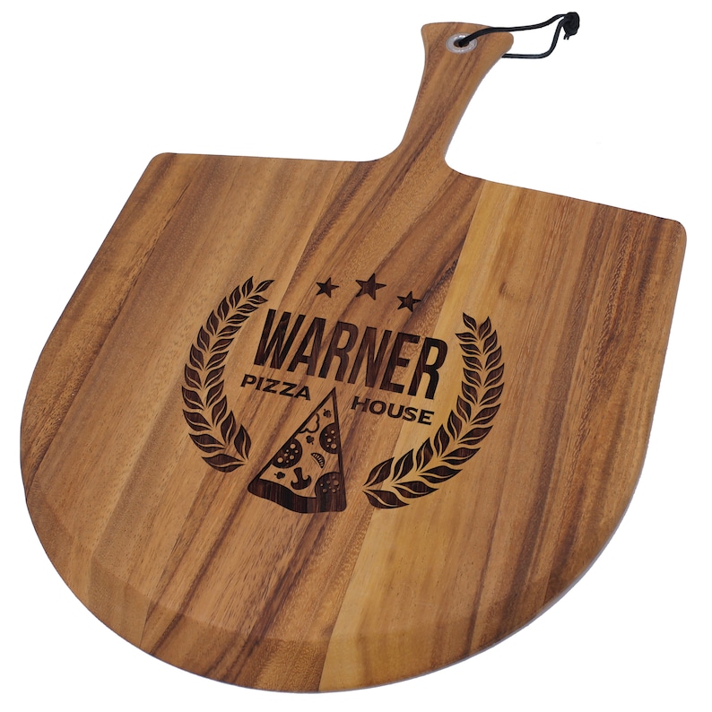 Peut inclure: Une pelle &agrave; pizza en bois avec une poign&eacute;e et un cordon noir pour suspendre. La pelle a un bord incurv&eacute; et pr&eacute;sente le texte "WARNER PIZZA HOUSE" avec un dessin de part de pizza et une couronne de laurier.