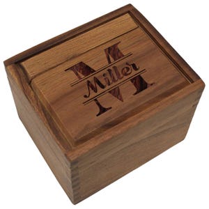May include: A wooden box with a sliding lid. The lid is engraved with the letter "M" and the name "Miller" in a decorative font. The box is made of a light brown wood with a darker wood inlay on the lid.