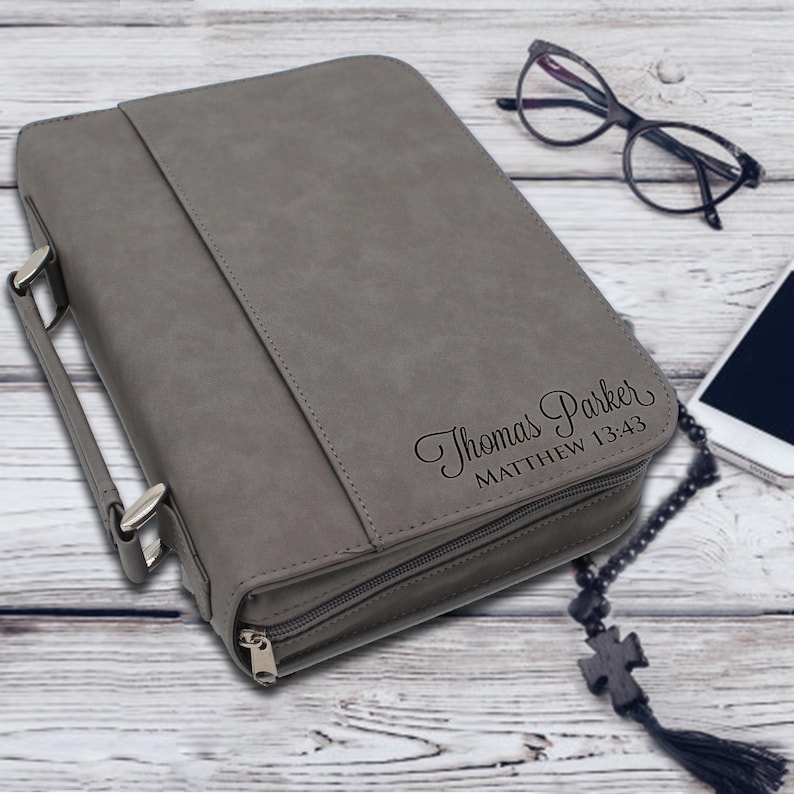 May include: Gray Bible cover with a handle and zipper closure. The cover has the name "Thomas Parker" and "Matthew 13:43" printed on it. A pair of glasses, a smartphone, and a rosary are next to the Bible cover.