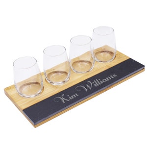 May include: A wooden serving tray with four stemless wine glasses. The tray has a bamboo base with a black slate insert engraved with the name "Kim Williams". The glasses are clear and empty, ready for use.