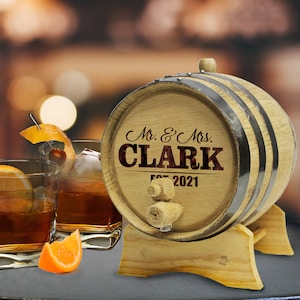 May include: A wooden barrel with a metal band and a spigot. The barrel is engraved with the text "Mr. & Mrs. CLARK Est. 2021". The barrel is sitting on a wooden stand.
