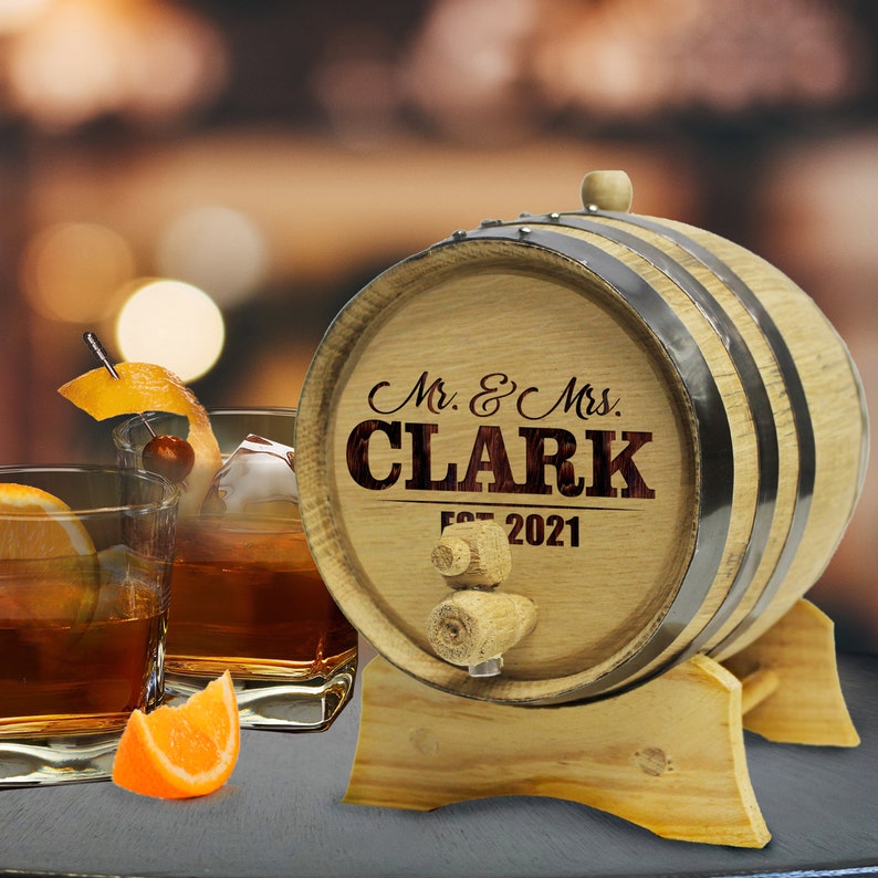 May include: A wooden barrel with a metal band and a spigot. The barrel is engraved with the text "Mr. & Mrs. CLARK Est. 2021". The barrel is sitting on a wooden stand.
