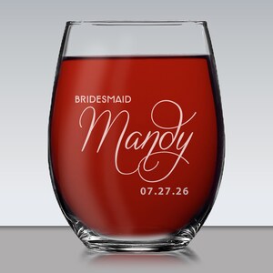 May include: A clear stemless wine glass filled with red liquid. The glass features white text that reads BRIDESMAID, the name Mandy in a large cursive script, and the date 07.27.26 printed in a clean sans-serif font on the front surface.