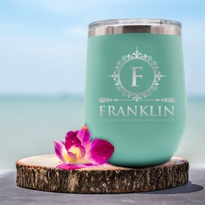 May include: A teal insulated tumbler with a silver lid, featuring the name "FRANKLIN" and the letter "F" in a decorative frame. A pink and purple orchid rests on a wooden slice in front of the tumbler. The background is a blurred beach scene.
