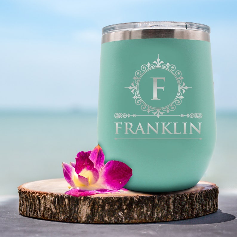 May include: A teal insulated tumbler with a silver lid, featuring the name "FRANKLIN" and the letter "F" in a decorative frame. A pink and purple orchid rests on a wooden slice in front of the tumbler. The background is a blurred beach scene.