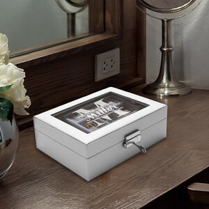 May include: A rectangular white jewelry box with a clear glass lid featuring the white printed text Miller and a large letter M. The box has a silver-toned metal latch on the front with a small key inserted, and the interior is divided into compartments.