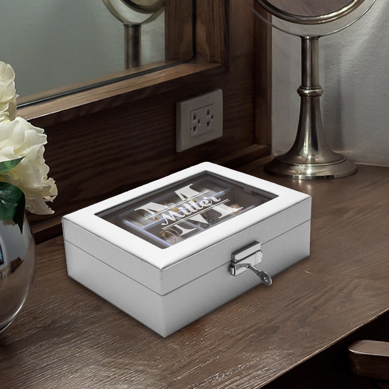 May include: A rectangular white jewelry box with a clear glass lid featuring the white printed text Miller and a large letter M. The box has a silver-toned metal latch on the front with a small key inserted, and the interior is divided into compartments.