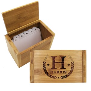 Puede incluir: Una caja de recetas rectangular de bamb&uacute; con tapa abatible que presenta un dise&ntilde;o grabado con l&aacute;ser de la letra H, el nombre HARRIS, dos estrellas y una corona de laurel. La caja abierta contiene fichas blancas con pesta&ntilde;as de la A a la E.