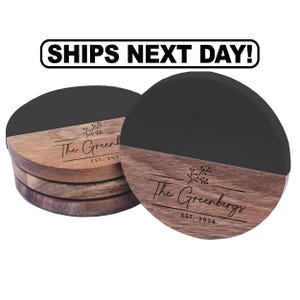 May include: A stack of round coasters featuring a two-tone design with a matte black upper section and a natural brown wood grain lower section. Each coaster is engraved with a floral branch icon, the text The Greenbergs, and EST. 2026.