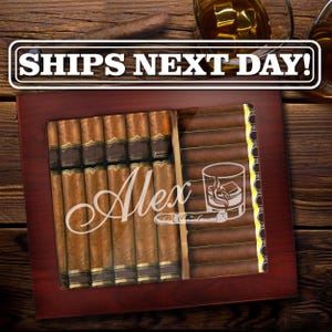 May include: A dark wood cigar humidor with a glass top featuring the name Alex in white script, a graphic of a glass of whiskey with ice, and a cigar. The humidor is filled with rows of brown cigars with gold bands, and the text SHIPS NEXT DAY!