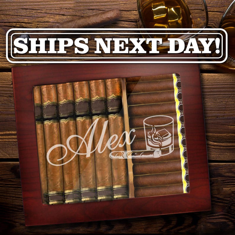 May include: A dark wood cigar humidor with a glass top featuring the name Alex in white script, a graphic of a glass of whiskey with ice, and a cigar. The humidor is filled with rows of brown cigars with gold bands, and the text SHIPS NEXT DAY!