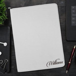 May include: A rectangular white leather-style portfolio case with a zip closure along the top edge. The front surface features the name Williams embossed in a dark, elegant script font in the bottom right corner with visible stitching.