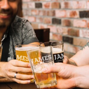 May include: Three clear beer glasses are raised in a toast. The glasses are filled with light and dark beers. The glasses have the words "JAMES JAN" and "Groomsmen" printed on them. The background is a brick wall.