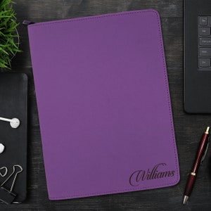 May include: A rectangular purple portfolio folder with a zipper closure along the top edge. The front cover features the name Williams engraved in a dark cursive script in the bottom right corner, with visible stitching along the perimeter.