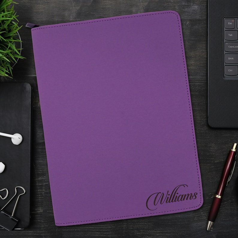 May include: A rectangular purple portfolio folder with a zipper closure along the top edge. The front cover features the name Williams engraved in a dark cursive script in the bottom right corner, with visible stitching along the perimeter.