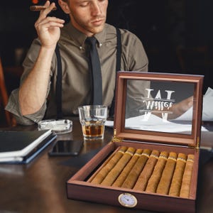 May include: A wooden cigar humidor with a glass lid featuring the white text Miller and a monogram M. The humidor contains eight brown cigars and has a round gold-rimmed analog hygrometer on the front exterior panel of the dark wood box.