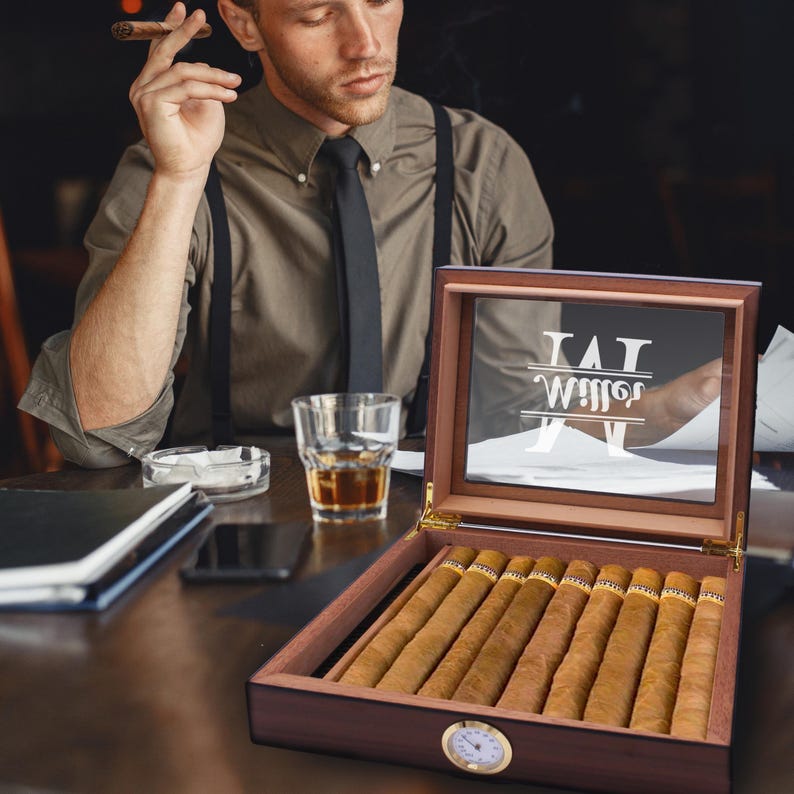 May include: A wooden cigar humidor with a glass lid featuring the white text Miller and a monogram M. The humidor contains eight brown cigars and has a round gold-rimmed analog hygrometer on the front exterior panel of the dark wood box.