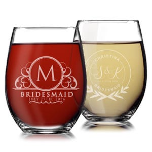 May include: Two stemless wine glasses with etched designs. The left glass contains red liquid and features a letter M inside a circle with the text BRIDESMAID and JULY 27TH, 2026. The right glass contains white liquid with a circular design, the name CHRISTINA, the initials S&K, and the text JULY 27TH, 2026 and BRIDESMAID.