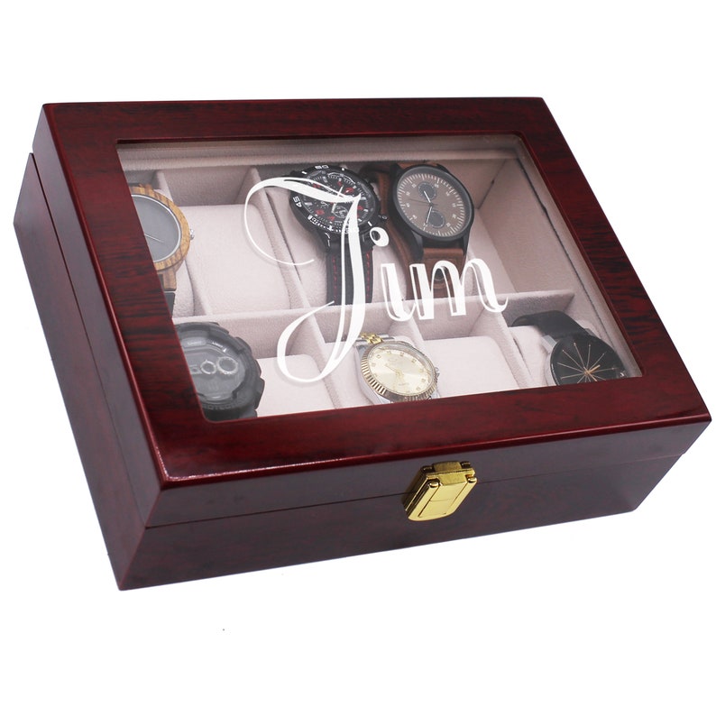 May include: A wooden watch box with a clear glass top, displaying several watches. The box is a dark reddish-brown color with a gold clasp. The name "Jim" is etched on the glass in a cursive font.