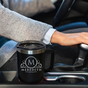 Pode incluir: Caneca de viagem preta com aro e al&ccedil;a prateados. A caneca apresenta um monograma com a letra "M" dentro de um c&iacute;rculo decorativo e o nome "Meredith" abaixo. A caneca est&aacute; em um carro.