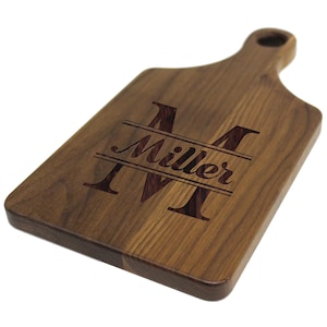 May include: A personalized walnut wood cutting board with a large "M" initial and the name "Miller" engraved in a script font.