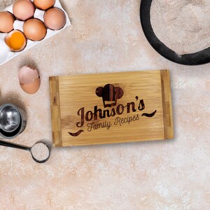 Puede incluir: Una tabla de cortar rectangular de bamb&uacute; con un icono de gorro de chef grabado a l&aacute;ser y el texto "Johnson's Family Recipes" en el centro. La tabla est&aacute; rodeada de huevos marrones en un cart&oacute;n, una c&aacute;scara de huevo, harina y una cuchara.