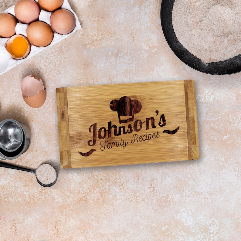 Puede incluir: Una tabla de cortar rectangular de bamb&uacute; con un icono de gorro de chef grabado a l&aacute;ser y el texto "Johnson's Family Recipes" en el centro. La tabla est&aacute; rodeada de huevos marrones en un cart&oacute;n, una c&aacute;scara de huevo, harina y una cuchara.