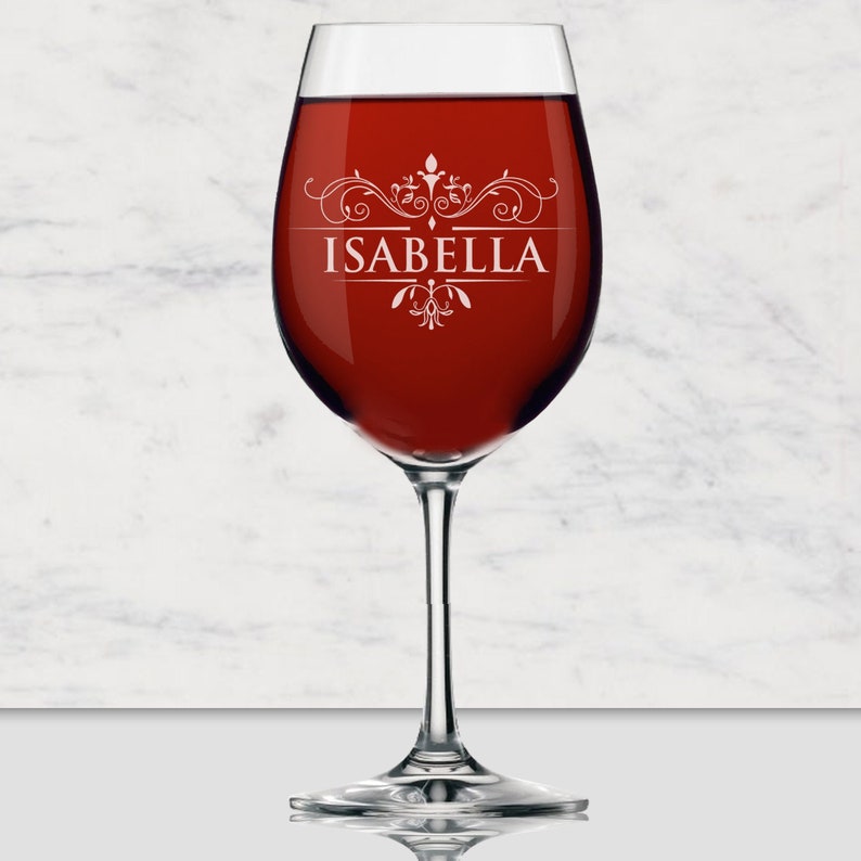 May include: A clear glass wine glass filled with deep red wine. The glass features a white etched design with decorative scrollwork and the name ISABELLA centered in capital letters. The stem and base are transparent glass with a smooth finish.