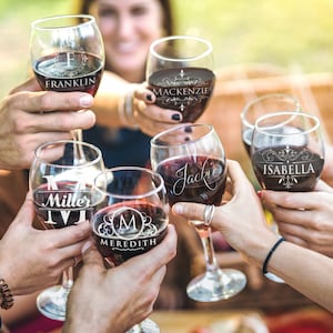 May include: Six clear glass wine glasses filled with red wine, each featuring a unique white etched design. The glasses are personalized with the names Franklin, Mackenzie, Miller, Jackie, Meredith, and Isabella, along with decorative monograms.