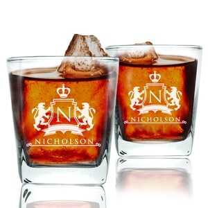Op de afbeelding: Twee heldere glazen tumblers met een gepersonaliseerd ontwerp. Elk glas heeft een wit wapenschild met een kroon, de letter "N" en twee leeuwen. De tekst "Nicholson" staat onder het wapenschild.