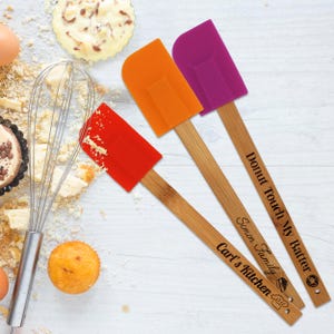 May include: Three silicone spatulas with wooden handles. The spatula heads are red, orange, and purple. The wooden handles have text that reads "Carl's Kitchen", "Simon Family", and "Donut Touch My Batter". Baking-themed.