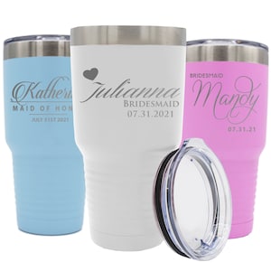 May include: Three stainless steel tumblers with lids. The tumbler on the left is light blue with the text "Katherin Maid of Honor July 31st 2021". The tumbler in the center is white with the text "Julianna Bridesmaid 07.31.2021". The tumbler on the right is pink with the text "Bridesmaid Mandy 07.31.21".