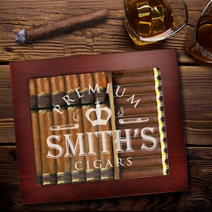 May include: A wooden humidor with a glass top, filled with cigars. The humidor has the text "PREMIUM SMITH'S CIGARS" in white. A lit cigar and two glasses of whiskey are on a wooden surface.