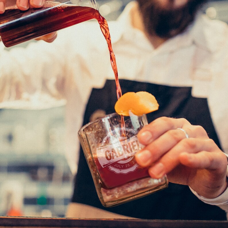 Peut inclure: Une personne verse un liquide rouge d'un pichet en verre dans un verre transparent avec une &eacute;tiquette personnalis&eacute;e qui dit "Gabriel" et "Juillet 25, 20".