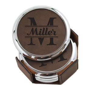 May include: Set of four brown leather coasters with a silver metal rim. The coasters feature a personalized monogram with the initial "M" and the name "Miller" in a split monogram style. The coasters are in a brown leather holder.
