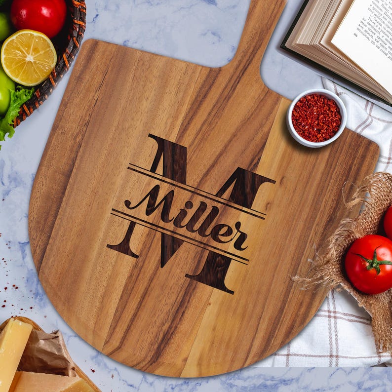 Peut inclure: Une pelle &agrave; pizza en bois avec une poign&eacute;e, portant le nom "Miller" grav&eacute; dans une police d&eacute;corative &agrave; l'int&eacute;rieur d'un grand "M". La pelle est entour&eacute;e d'ingr&eacute;dients comme des tomates, des &eacute;pices et du fromage.