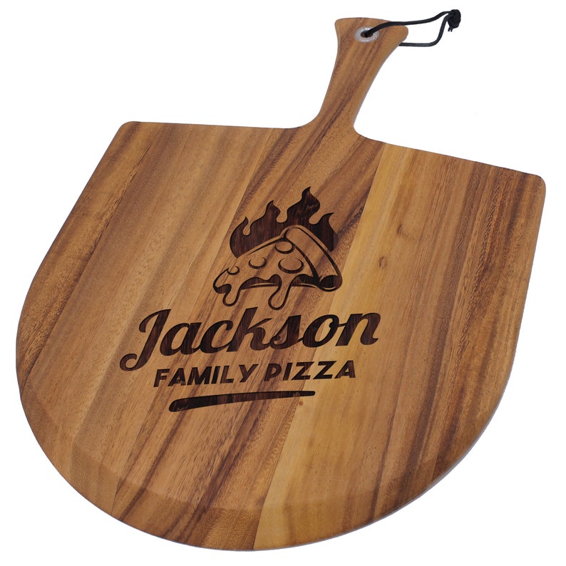 Peut inclure: Une pelle &agrave; pizza en bois avec une poign&eacute;e et une lame incurv&eacute;e. La pelle pr&eacute;sente le texte grav&eacute; "Jackson FAMILY PIZZA" avec un motif de part de pizza et de flammes. Un cordon noir est attach&eacute; &agrave; la poign&eacute;e.