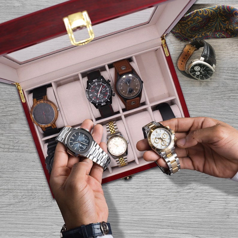 May include: A red wooden watch case with a glass top, filled with various wristwatches. Two watches are held in hands. The watches have black, brown, and silver bands and faces. A patterned tie is visible.