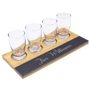 May include: A wooden shot glass flight set with four clear glasses. The set includes a bamboo base with a dark gray slate insert engraved with the name "Jim Williams".