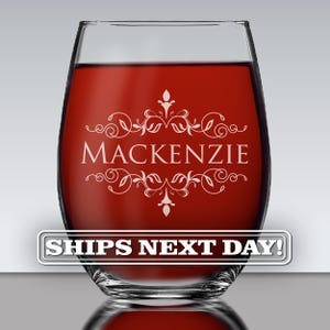 May include: A clear stemless wine glass filled with red liquid. The glass features an etched design with the name MACKENZIE centered between two symmetrical, ornate floral flourishes. The glass has a smooth, rounded silhouette and a thick base.