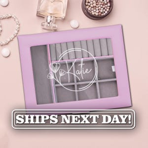 May include: A rectangular light purple jewelry box with a clear glass lid featuring a white circular graphic with the name Katie and a floral design. The interior is lined with grey velvet and divided into multiple compartments for storage.