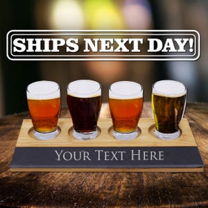 May include: A rectangular bamboo and slate beer flight board featuring four circular cutouts holding four clear glass beer sampler glasses filled with different colored beers. The slate section at the front displays the text YOUR TEXT HERE.