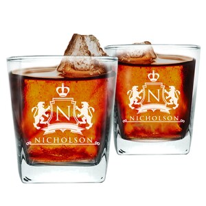 Może przedstawiać: Dwa przezroczyste szklane kieliszki do whisky z personalizowanym wzorem. Każdy kieliszek ma herb z koroną, literą "N" i dwoma lwami. Tekst "NICHOLSON" jest wygrawerowany pod herbem.
