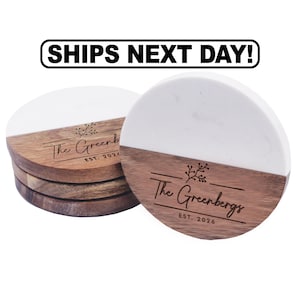May include: A stack of round coasters featuring a split design of white marble and brown wood. The wood section is engraved with a floral graphic, the text The Greenbergs, and the text EST. 2026. Above the coasters is the text SHIPS NEXT DAY!