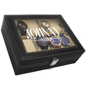 May include: A black watch display case with a glass top, holding several watches. The case is rectangular, with white stitching and a silver clasp. The word "JOHNNY" is printed on the glass. The watches have various band and face colors.