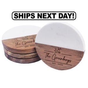 May include: A stack of round coasters featuring a split design of white marble and brown wood. The wood section is engraved with a floral graphic, the text The Greenbergs, and the text EST. 2026. Above the coasters is the text SHIPS NEXT DAY!