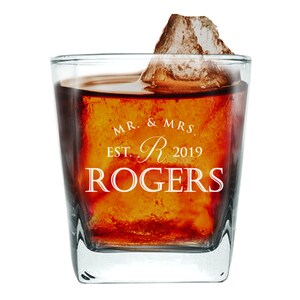 May include: A square, clear glass with a thick base, filled with a dark amber liquid and a large ice cube. The glass is engraved with the text "MR. & MRS. EST. R 2019 ROGERS".
